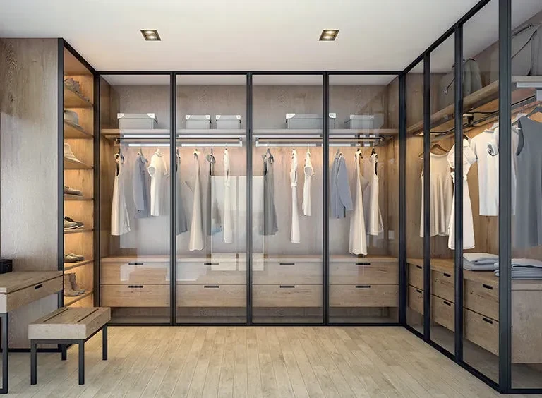 GEORGE L-shaped wardrobe design with dark framed transparent glass doors, lit hanging zones and drawers.
